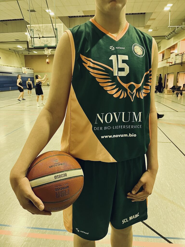 Basketballspieler steht in der Basktballhalle. Er hält einen Basketball unter dem rechten Arm und trägt ein Grün-Oranges Basketballtrikot mit der Spielernummer 15. Aufgedruckt ist der Sponsor des Teams: NOVUM – Der Biolierferservice.