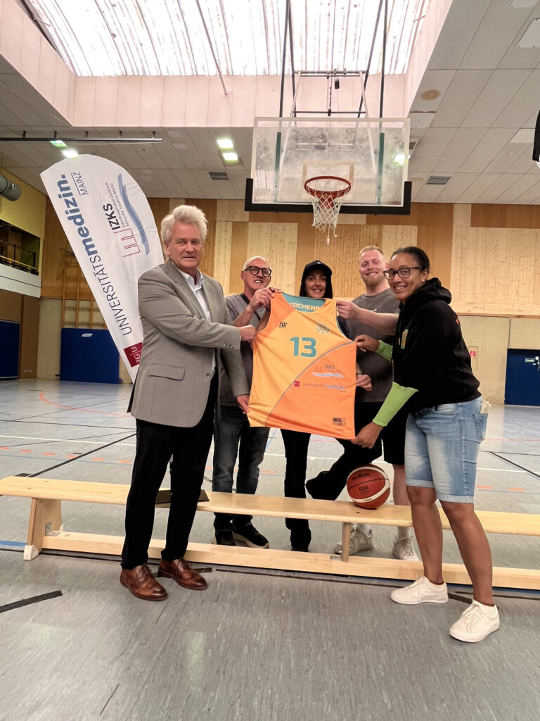 Fünf Personen halten ein Baskteballtrikot zwischen sich hoch. Es handelt sich um die Trikotübergabe mit dem Sponsor IZKS Mainz.